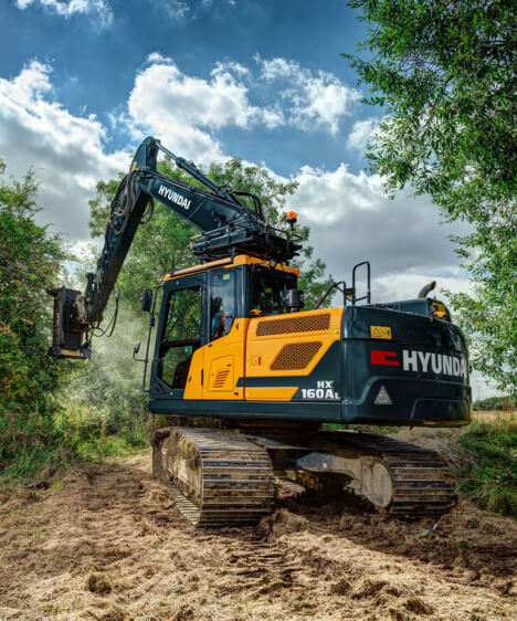 HD Hyundai HX160A L crawler excavator in a forest using the hedge trimmer attachment