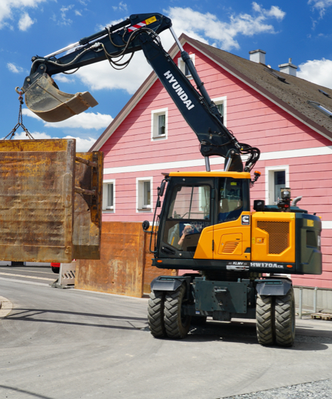 Excavatrice HD Hyundai HW170 CR déplaçant un gros objet en fer à côté d'une maison rose